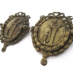 May include: Two antique brass door knockers with a decorative design featuring a woman standing next to a column. The knockers are oval shaped and have a wreath-like design around the edges.