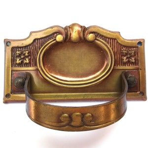 Può includere: Una maniglia per porta in ottone vintage con un design ornamentale. La maniglia ha una base rettangolare con un centro ovale rialzato e un manico curvo.