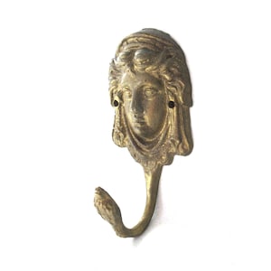 Op de afbeelding: Een gouden metalen haak met een decoratief gezichtsontwerp. Het gezicht is gebeeldhouwd met gedetailleerde kenmerken en een gedrapeerde hoofdtooi. De haak is in vintage stijl en zou een unieke toevoeging zijn aan een interieurproject.