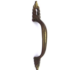 Puede incluir: Un tirador de puerta antiguo de latón con un diseño ornamentado. El tirador presenta una forma curvada con detalles decorativos de hojas y cuerda. El tirador es de un color latón cálido y envejecido, lo que sugiere encanto y artesanía vintage.