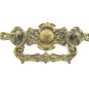 Peut inclure: Une poignée de tiroir en laiton ornée avec un motif central décoratif et deux boutons. La poignée a un design enroulé et est de style vintage.