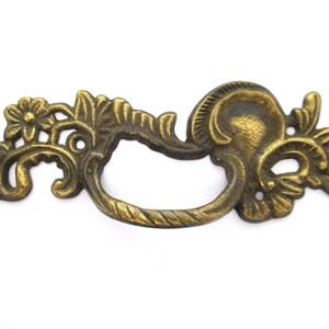 Könnte beinhalten: Verzierter, goldfarbener Metall-Schubladengriff mit floralem Design. Der Griff hat eine geschwungene Form und eine seilartige Textur.