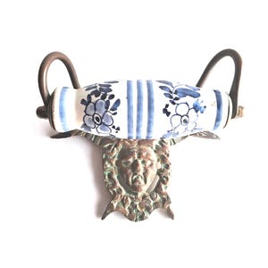 May include: Antique toilet paper holder with a ceramic roller featuring blue floral and striped patterns. The holder has bronze-colored metal accents and a decorative face design.