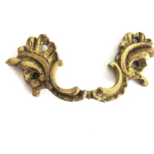 May include: A gold-toned metal decorative drawer pull with a floral design. The pull has two holes for mounting.
