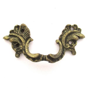 Puede incluir: Un tirador de cajón de metal dorado con un diseño decorativo y ornamentado. El tirador tiene forma de U con intrincados adornos en cada lado.