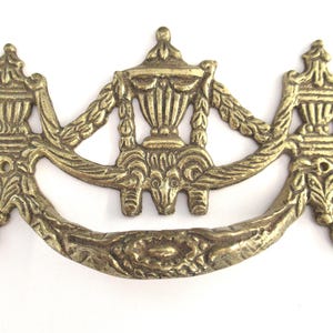 Könnte beinhalten: Verzierter Messing-Schubladengriff mit einem dekorativen Design, das eine zentrale Urne und florale Motive zeigt. Der Griff hat eine Halbmondform und eine glatte, polierte Oberfläche.