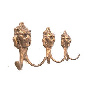 Op de afbeelding: Een wandkapstok met drie messingkleurige metalen haken, elk bekroond met een gebeeldhouwde leeuwenkop. De haken zijn van links naar rechts in afnemende grootte gerangschikt, waarbij elke leeuwenkop gedetailleerde gelaatstrekken vertoont.