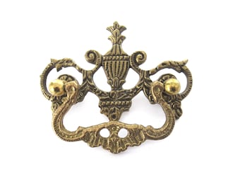 May include: Ornate brass cabinet pull with a decorative design featuring a central urn and two scroll-like handles. The pull has a gold finish and is accented with small round knobs.