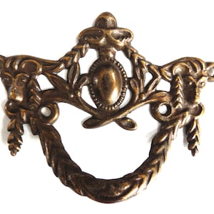 Op de afbeelding: Een decoratieve messing ladegreep met een bloemmotief en een centrale ovale vorm. De greep heeft een ringvormige handgreep en twee kleine gaten aan beide zijden.