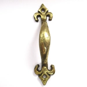 Könnte beinhalten: Ein Vintage-Messinggriff mit einem Fleur-de-Lis-Design an jedem Ende. Der Griff hat eine gebogene, ovale Mitte zum Greifen. Das Messing hat ein leicht gealtertes, angelaufenes Aussehen.
