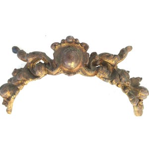 May include: A gold-toned metal decorative piece with a central oval design and intricate scrollwork on either side. The piece is likely a decorative element for furniture or a mirror.