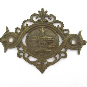 May include: A gold-toned metal decorative piece with an ornate design. The centre features a circular medallion with a crown and other decorative elements.