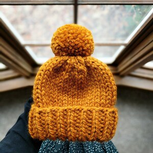 May include: A knitted beanie hat in a mustard yellow color with a fluffy pom pom on top.