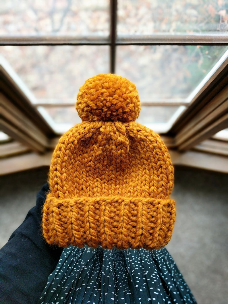 May include: A knitted beanie hat in a mustard yellow color with a fluffy pom pom on top.
