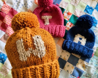 May include: Three chunky knit beanies with pom-poms on top. The mustard yellow hat features a white letter H, the pink hat features a white letter T, and the navy blue hat features a white letter A. Each hat has a thick ribbed cuff.