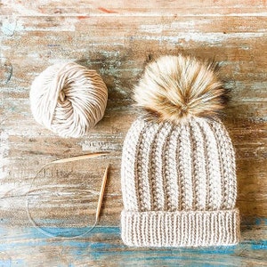May include: A beige knitted beanie with a fluffy brown pom pom, a ball of beige yarn, and two wooden knitting needles.