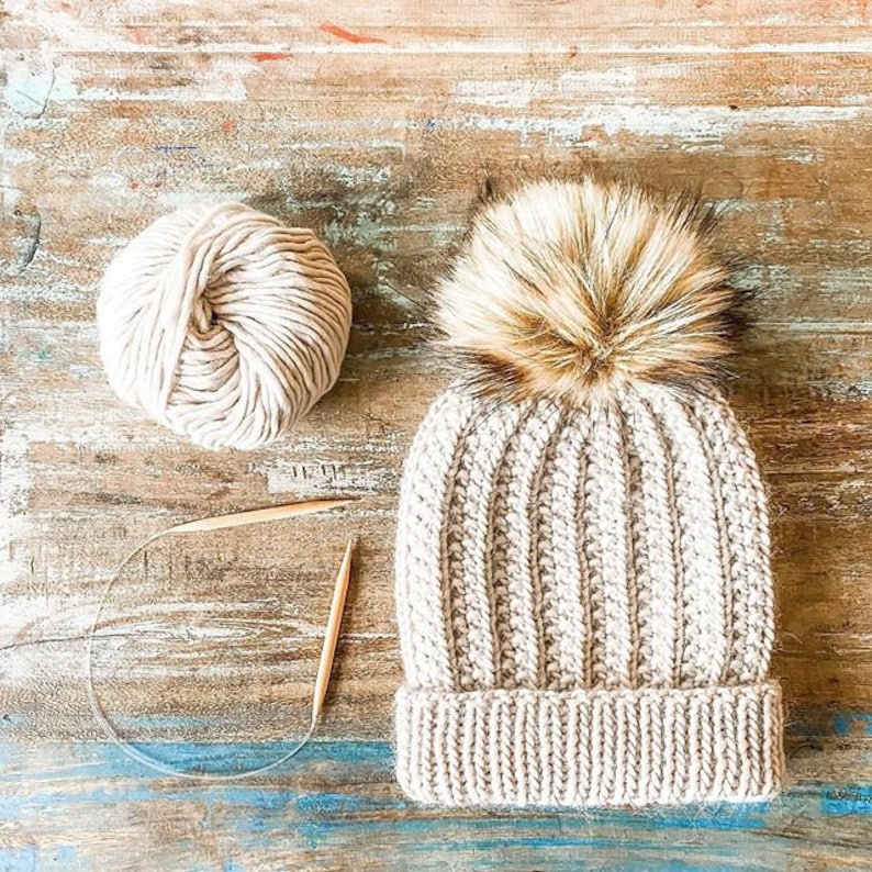May include: A beige knitted beanie with a fluffy brown pom pom, a ball of beige yarn, and two wooden knitting needles.
