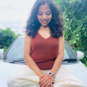 Peut inclure: Une femme portant un haut en crochet rouille et un pantalon blanc est assise sur le capot d'une voiture blanche. Elle a de longs cheveux noirs et porte une montre argent&eacute;e au poignet gauche.