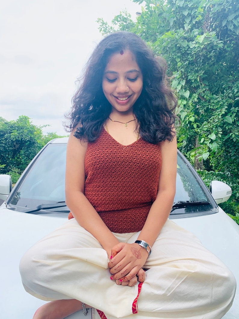 Peut inclure: Une femme portant un haut en crochet rouille et un pantalon blanc est assise sur le capot d'une voiture blanche. Elle a de longs cheveux noirs et porte une montre argent&eacute;e au poignet gauche.