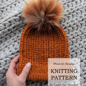May include: A knitted beanie in a burnt orange color with a fluffy brown and orange pom pom on top. The beanie is on a gray knitted surface. The text "Phoenix Beanie KNITTING PATTERN By Lady Jay Crochet" is on the image.