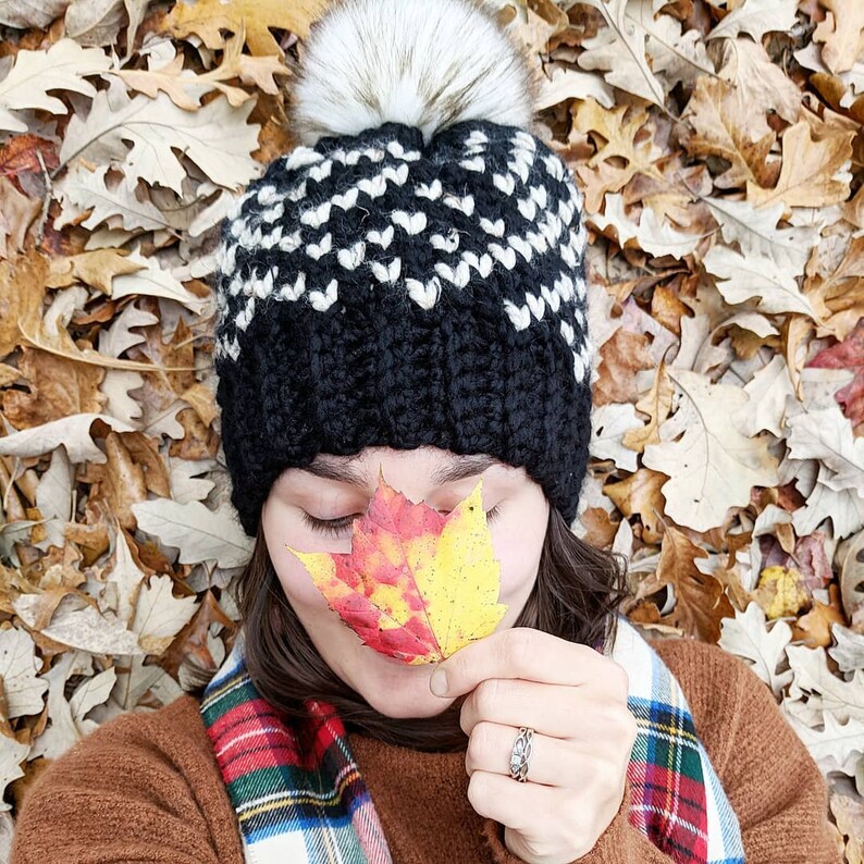 May include: A person wearing a black and white knit hat with a white pom pom, a red and black plaid scarf, and a brown sweater. They are holding a red and yellow leaf in front of their face. The background is a pile of brown leaves.