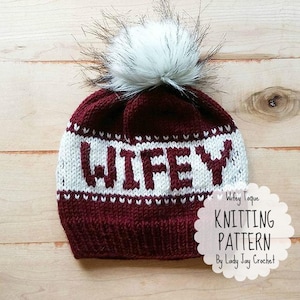May include: A burgundy and white knit beanie with the word "WIFEY" in bold letters. The hat features a white faux fur pom-pom on top. The design includes a knitting pattern by Lady Jay Crochet.
