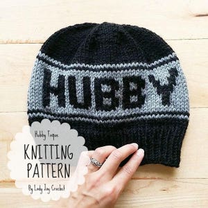 May include: Black and grey knitted toque with the word "HUBBY" knitted into the brim. The toque is on a wooden surface.