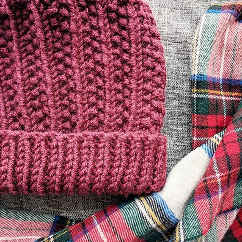 May include: A burgundy crocheted beanie hat with a textured stitch pattern. The hat is lying on a plaid fabric background.