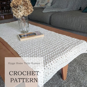 May include: A white crocheted table runner with a subtle textured pattern. The runner is on a wooden coffee table with a book and a vase of dried flowers. The text "Hygge Home Table Runner CROCHET PATTERN By Lady Jay Crochet" is on the image.