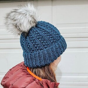 May include: A blue knitted beanie with a fluffy white pom pom. The beanie is made with a textured stitch pattern.