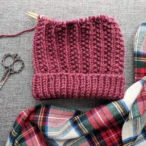 May include: A maroon knitted beanie hat with a ribbed texture, lying on a plaid fabric. The hat is partially finished, with a wooden knitting needle sticking out of the top.