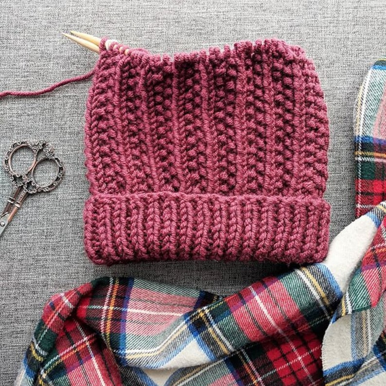 May include: A maroon knitted beanie hat with a ribbed texture, lying on a plaid fabric. The hat is partially finished, with a wooden knitting needle sticking out of the top.
