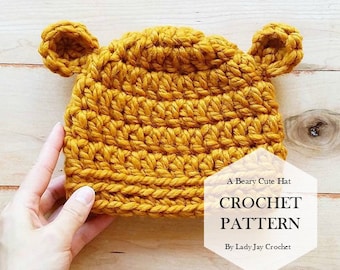 May include: A yellow crocheted hat with bear ears. The hat is on a wooden surface. The text "A Beary Cute Hat CROCHET PATTERN By Lady Jay Crochet" is on the image.