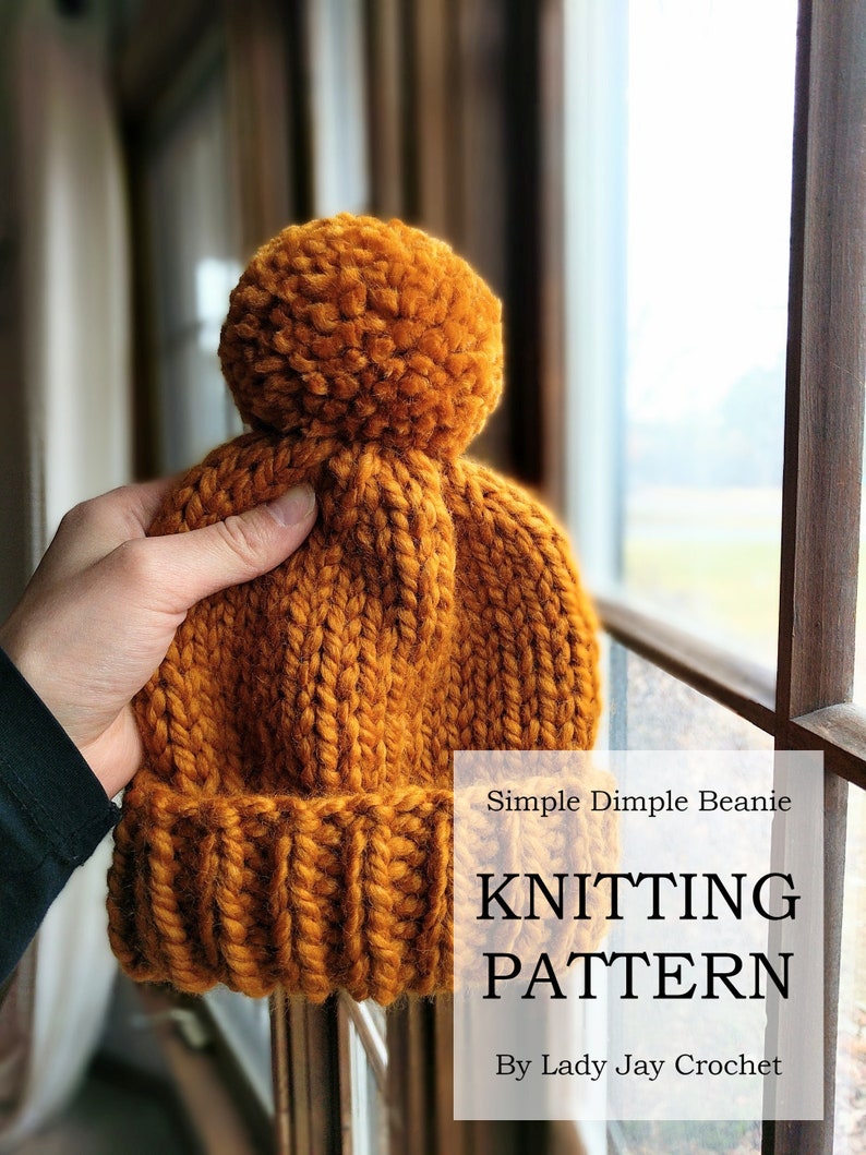 May include: A knitted beanie with a pom pom in a mustard yellow color. The beanie is held in a hand and the text "Simple Dimple Beanie Knitting Pattern By Lady Jay Crochet" is visible below the beanie.