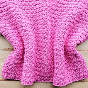 Peut inclure: Une couverture en crochet rose avec une surface textur&eacute;e. La couverture est pli&eacute;e et drap&eacute;e sur une surface en bois.