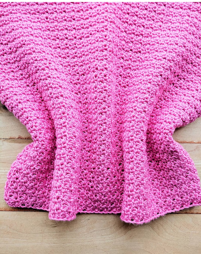 Peut inclure: Une couverture en crochet rose avec une surface textur&eacute;e. La couverture est pli&eacute;e et drap&eacute;e sur une surface en bois.
