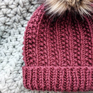 May include: A burgundy knitted beanie with a fluffy brown pom pom on top. The beanie is made with a textured stitch pattern.