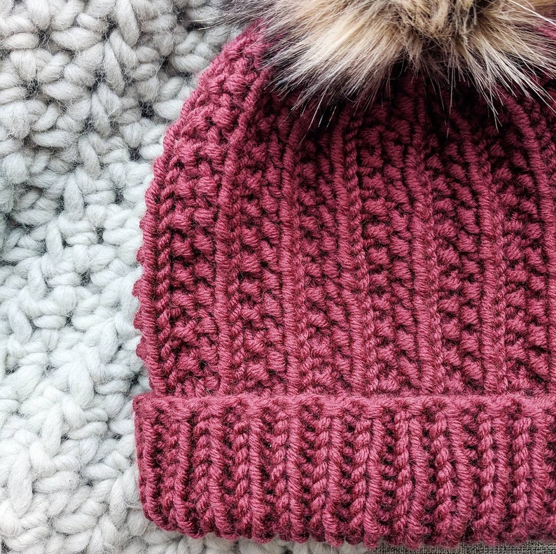 May include: A burgundy knitted beanie with a fluffy brown pom pom on top. The beanie is made with a textured stitch pattern.