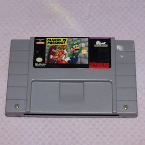 May include: A Super Nintendo Entertainment System (SNES) game cartridge for the game "Mario is Missing!"  The cartridge is gray with a label featuring a colorful image of Mario, Luigi, and Toad. The label also includes the text "Licensed by Nintendo" and "Super Nintendo Entertainment System".