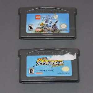 May include: Two Game Boy Advance game cartridges. The top cartridge is gray and features a blue and white label with the text "LEGO Knights Kingdom". The bottom cartridge is gray and features a blue and yellow label with the text "Island Xtreme Stunts".