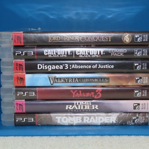 May include: A stack of six PlayStation 3 video game cases. The games are: Lord of the Rings Conquest, Call of Duty: Black Ops, Call of Duty: Black Ops II, Disgaea 3: Absence of Justice, Valkyria Chronicles, Yakuza 3, Tomb Raider: Underworld, and Tomb Raider.