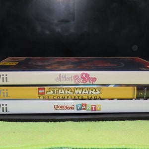 May include: A stack of three white Wii video game cases. The top case has a pink and white label with the text "Littlest Pet Shop". The middle case has a yellow label with the text "LEGO Star Wars The Complete Saga". The bottom case has a white label with the text "WordJong Party".