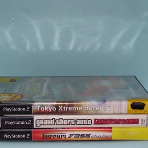 May include: Three PlayStation 2 video game cases stacked on top of each other. The games are "Tokyo Xtreme Racer 3", "Grand Theft Auto: Liberty City Stories", and "Ferrari F355 Challenge". The cases are black with colorful game art.