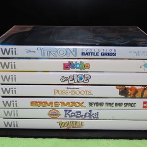 May include: A stack of seven Wii video game cases. Titles include "Tron: Evolution Battle Grids," "Elebits," "de Blob," "Puss in Boots," "Sam & Max Beyond Time and Space," "Kabookii," and "Thrillville: Off the Rails."