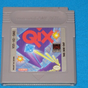 May include: A gray Game Boy cartridge with a red, white, and blue label. The label features the word "Qix" in large red letters and a blue diamond shape. The text "DMG-QX-USA" and "MADE IN JAPAN" is printed on the label.