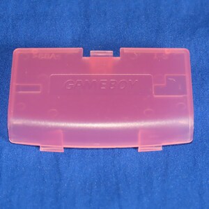May include: A translucent pink Game Boy battery cover. The word "GAMEBOY" is embossed on the cover. The cover has a rectangular shape with rounded edges and several tabs for attachment. The background is a blue fabric.