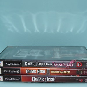 May include: Three PlayStation 2 video game cases stacked on top of each other. The top case is black with the text "Guitar Hero Encore Rocks the 80s". The middle case is orange with the text "Guitar Hero III Legends of Rock". The bottom case is red with the text "Guitar Hero".