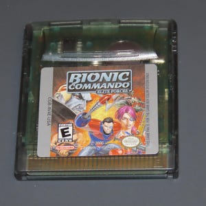 May include: A Bionic Commando: Elite Forces video game cartridge for the Game Boy Color. The cartridge is in a clear green plastic case. The label features a colorful illustration of characters and the game's title.