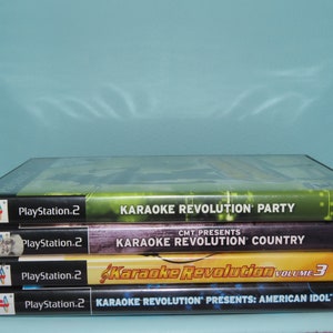 May include: Four PlayStation 2 game cases stacked on top of each other. The cases are for Karaoke Revolution Party, Karaoke Revolution Country, Karaoke Revolution Volume 3, and Karaoke Revolution Presents: American Idol.