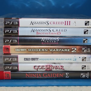 May include: A stack of eight video game cases for the Playstation 3 console. The games are Assassin's Creed III, Assassin's Creed Brotherhood, Assassin's Creed Revelations, Call of Duty: Modern Warfare 2, Call of Duty: Modern Warfare 3, God of War, and Ninja Gaiden Sigma.